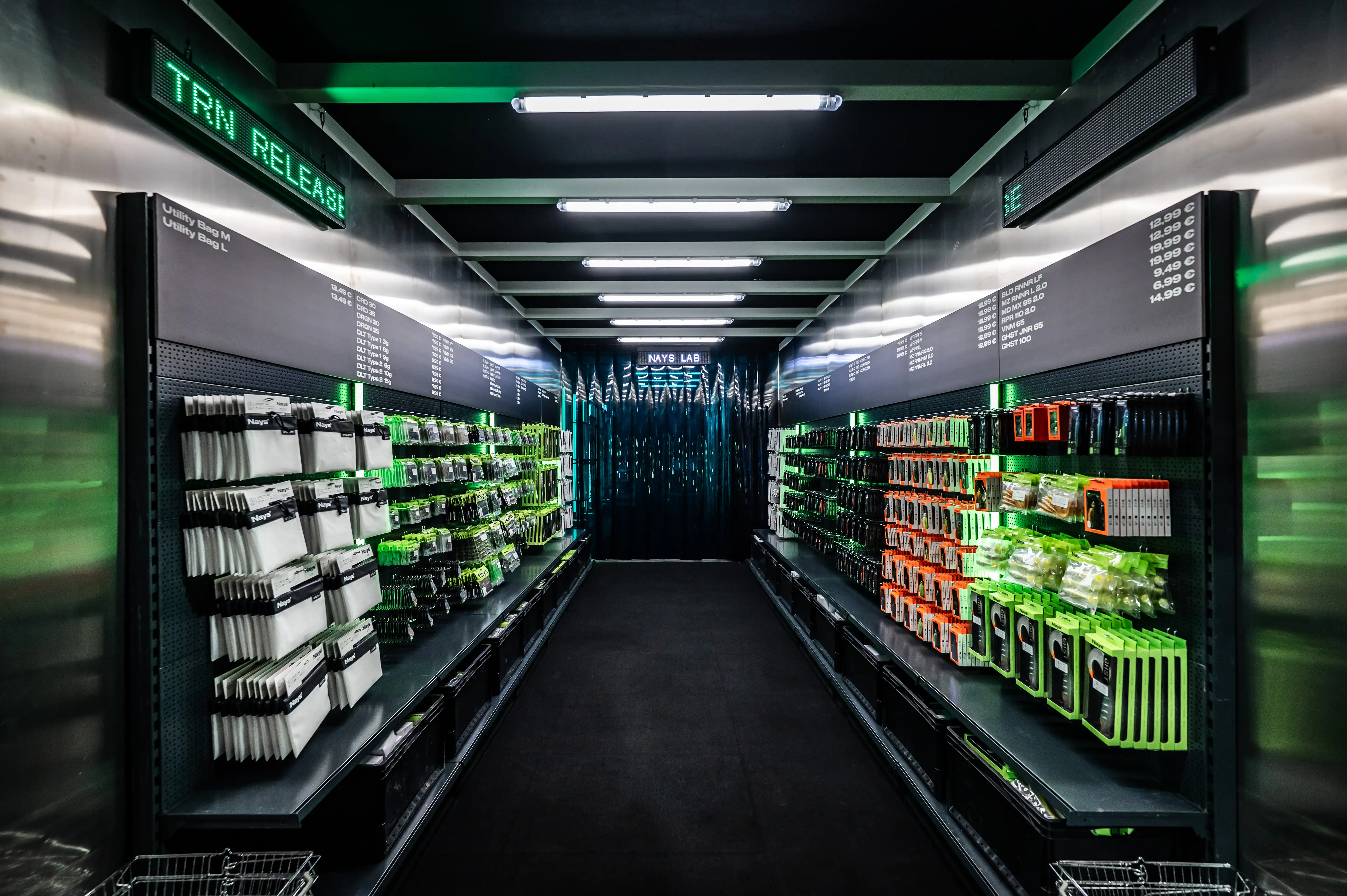 Dark supermarket-style aisle with green and white Nays lure packaging on black shelves under LED tickers.