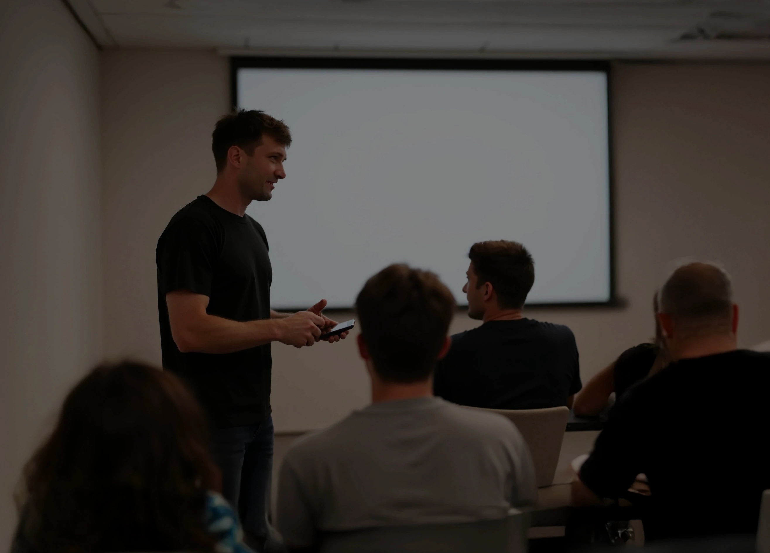 A person stands at the front of a classroom, engaging with seated attendees, with a blank projector screen in the background; the scene captures a learning or presentation environment.