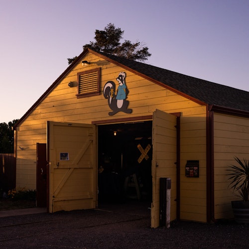 在黄昏时分，一座黄色的谷仓敞开着大门，外墙上画着一个卡通臭鼬，轮胎嵌在它的旁边。谷仓内可以看到一些工具和一个铁路道口标志。