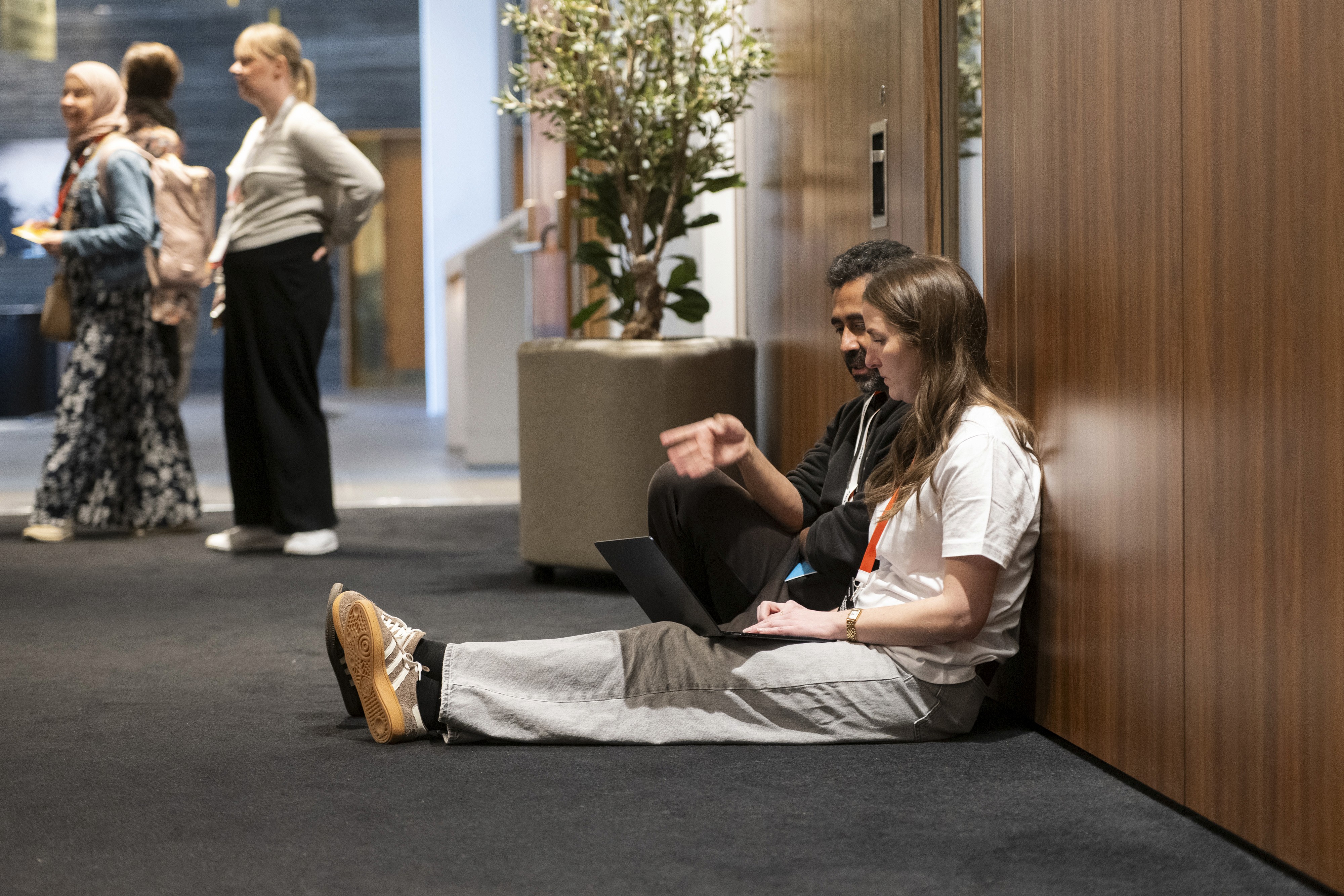 A person sits against a wall on the floor, using a laptop, while others stand and chat in the background.