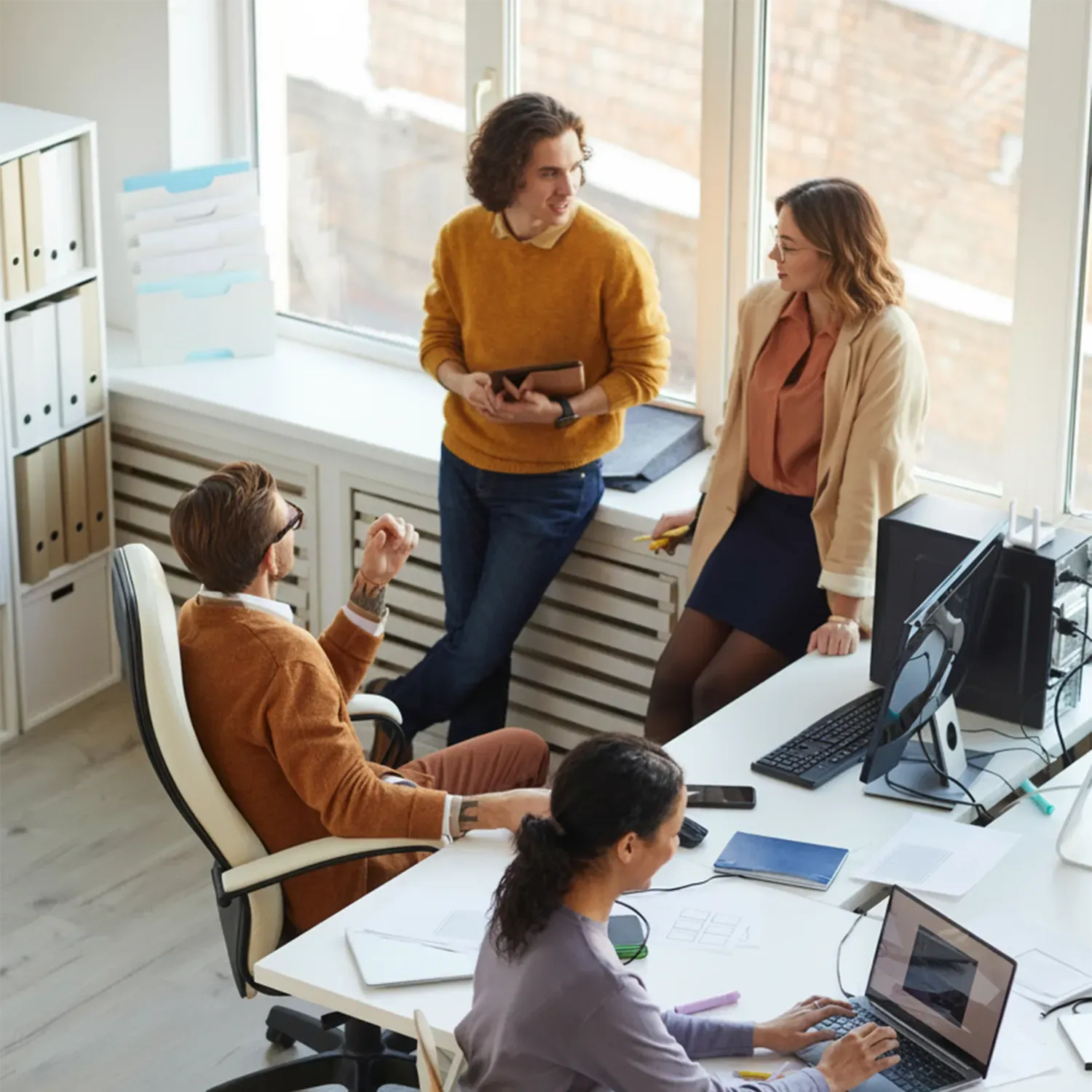 Skupina kolegů v kanceláři při neformální diskusi u stolu a okna, ilustrující týmovou spolupráci a komunikaci.