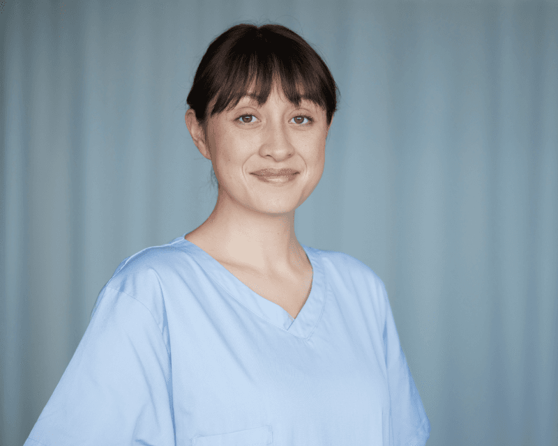 Portrait of a healthcare worker in light blue scrubs smiling against a soft, neutral backdrop.