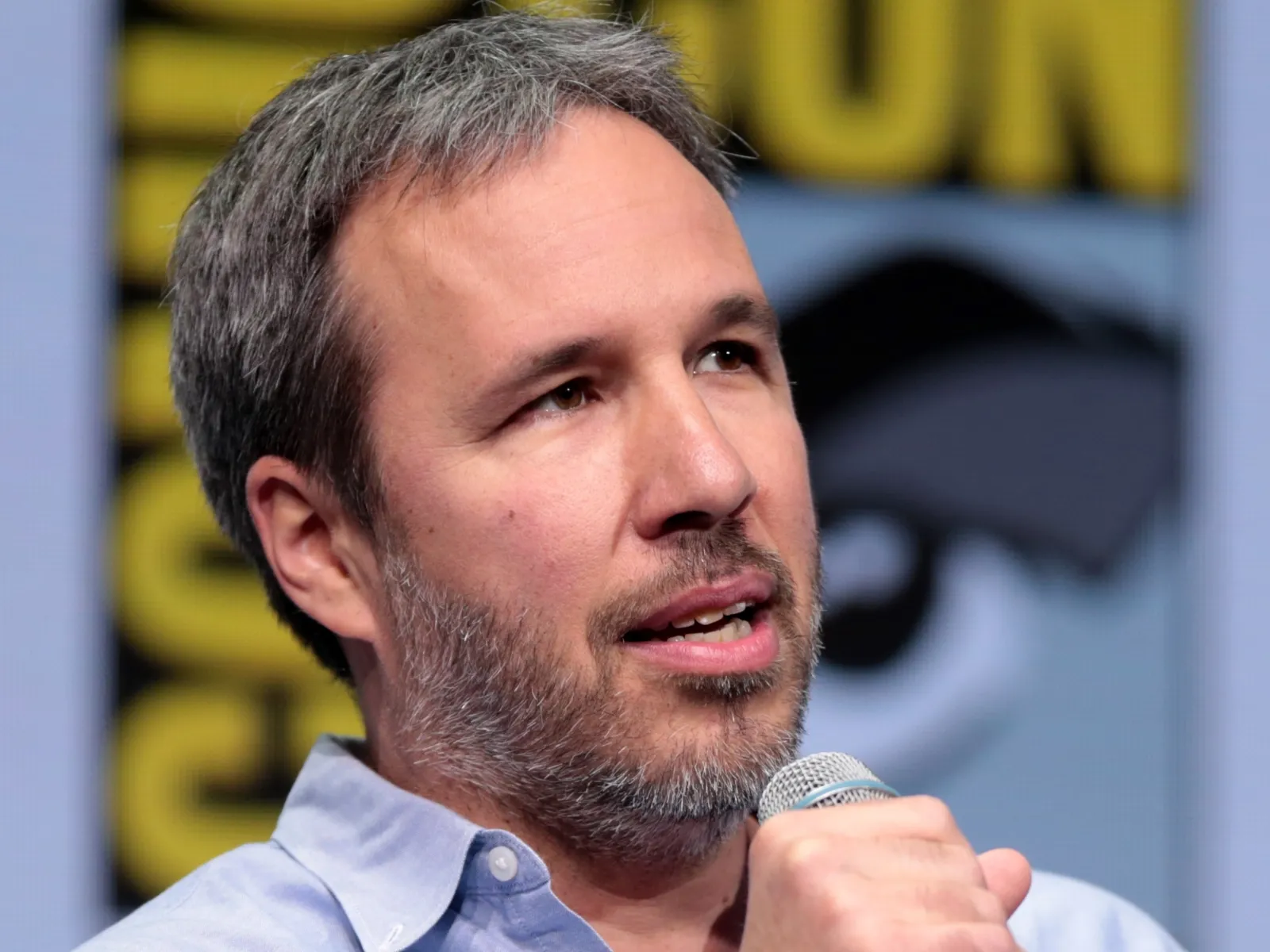 A man with a beard and grey hair speaks into a microphone at a panel event, with a blurred logo in the background.