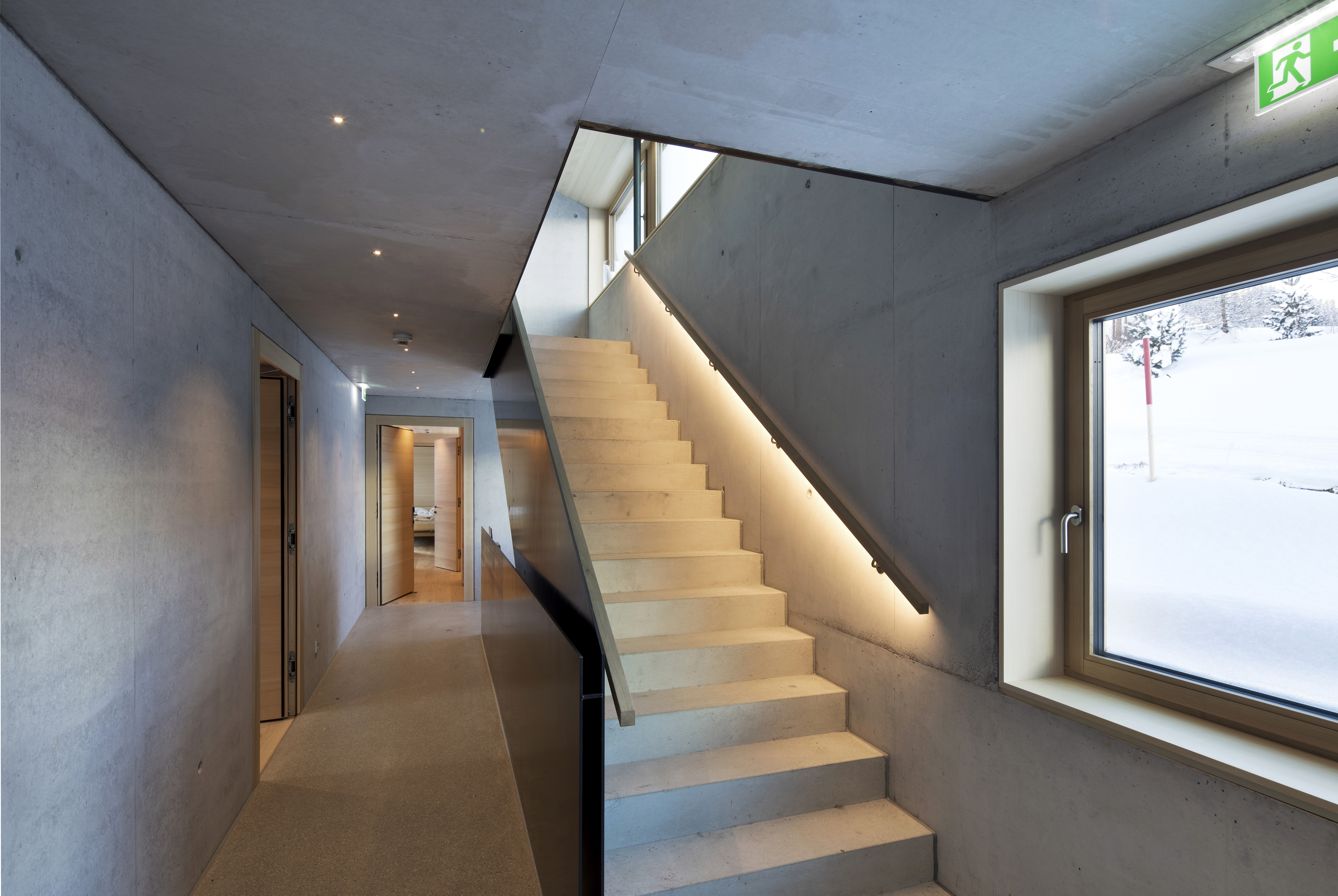 Modern concrete interior with a softly lit staircase, hallway with wooden doorframes, and a large window showing deep winter snow outside.