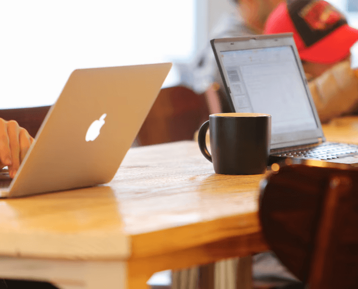 Two open laptops sit on a wooden table in a casual workspace, with a black coffee mug placed between them. One laptop shows a presentation screen, while a person nearby holds a reusable bottle. Another person is partially visible using a laptop with an illuminated logo. The background is softly blurred, suggesting a relaxed, collaborative café or meeting environment.