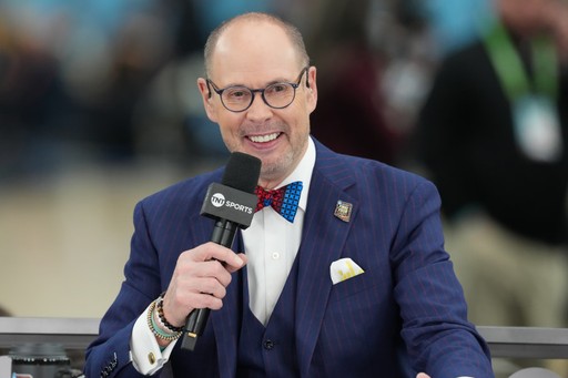 Ernie Johnson in a blue pinstripe suit, red and blue bow tie, and glasses holds a TNT Sports microphone while speaking during a live broadcast. A blurred audience is visible in the background.