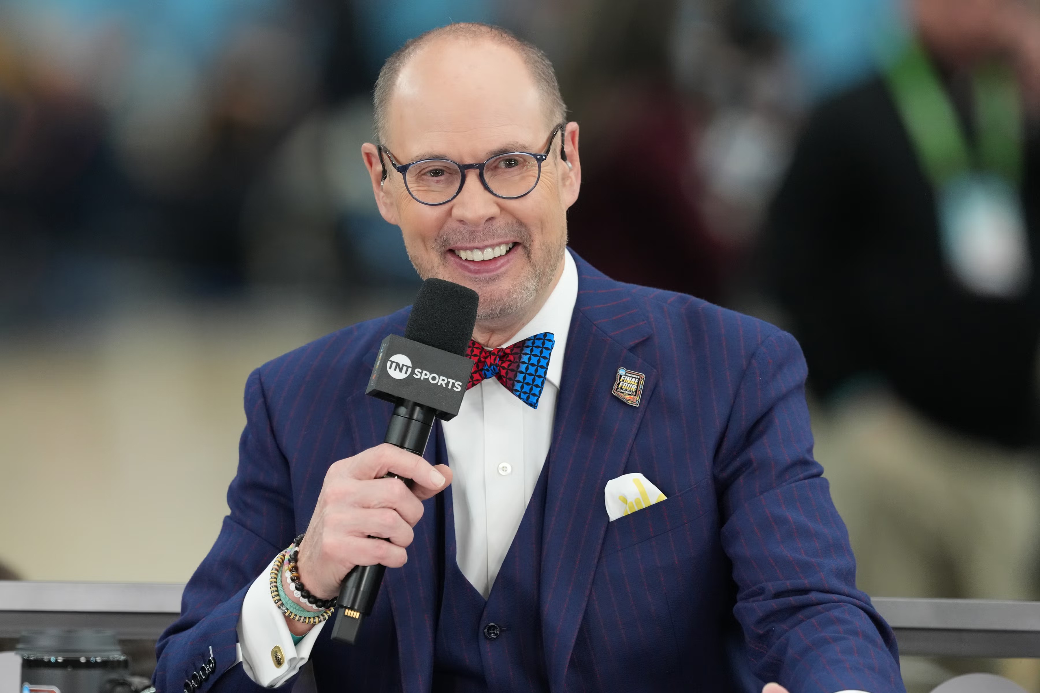 Ernie Johnson in a blue pinstripe suit, red and blue bow tie, and glasses holds a TNT Sports microphone while speaking during a live broadcast. A blurred audience is visible in the background.