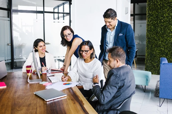 A imagem mostra um grupo de cinco pessoas em uma reunião de negócios em um escritório moderno. Eles estão reunidos ao redor de uma mesa de madeira, com laptops, papéis e gráficos à frente. Uma mulher ao centro parece estar explicando algo, enquanto os outros membros da equipe prestam atenção e discutem o que está sendo apresentado. A interação entre eles parece colaborativa, com todos envolvidos na discussão. O ambiente é moderno, com móveis confortáveis ao fundo e uma parede coberta de plantas, que adiciona um toque natural ao espaço. A imagem sugere um ambiente corporativo dinâmico e colaborativo.