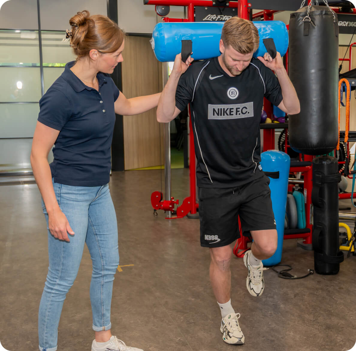 A woman in a navy polo shirt and jeans instructs a man in a black sports outfit holding a blue exercise bag over his shoulders, within a modern gym environment.