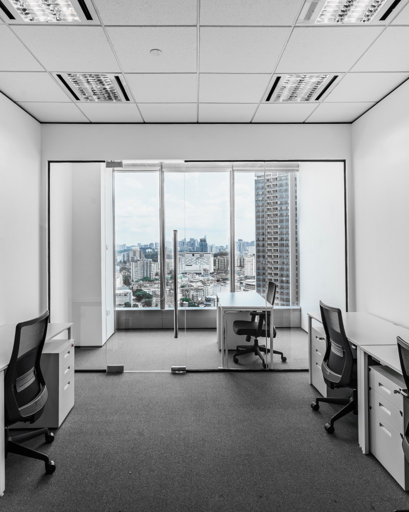 Inside a modern coworking space featuring minimalist design, with desks and chairs arranged to overlook a city skyline through large glass windows.