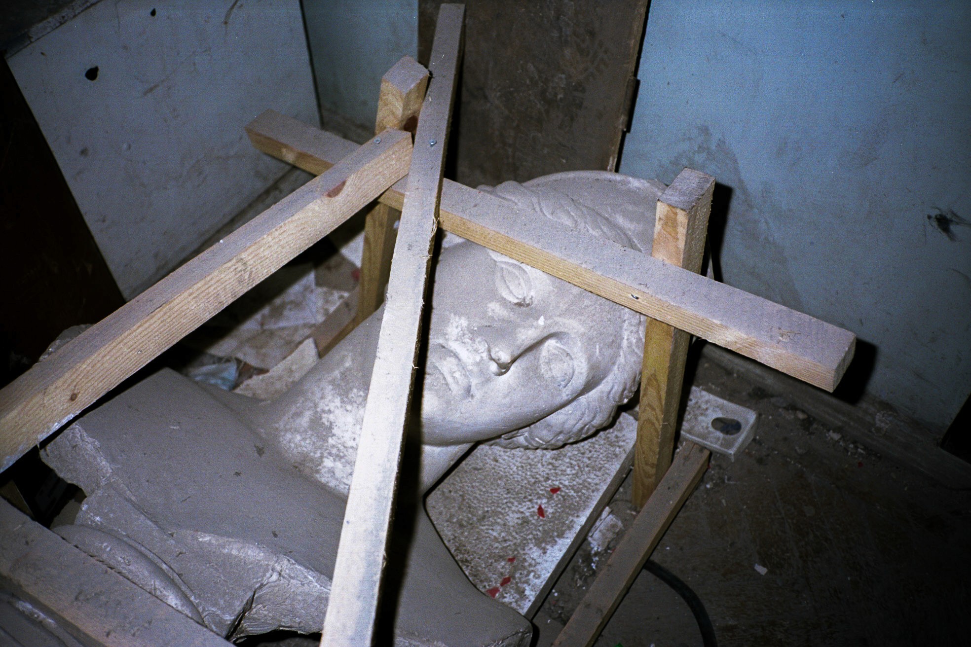 Broken plaster statue of a classical-style face lying on the floor, framed by wooden beams in a dimly lit, dusty room.