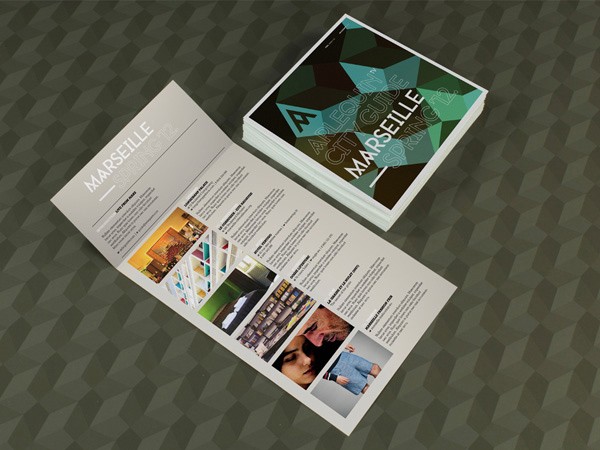 A tri-fold brochure lies open on a dark green geometric patterned surface. The brochure’s front panel displays a photograph of a building interior and text including “MARSEILLE,” “CITY GUIDE,” and “SUPER 12.” The back panel features smaller images and text, including “MARSEILLE” and “SUPER 12.” A stack of similar brochures is visible behind it.