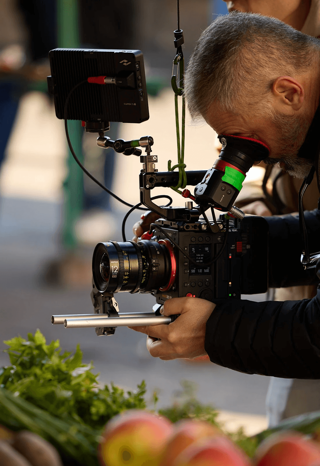 A person operates a professional film camera equipped with various attachments, capturing a scene in an outdoor setting surrounded by fruit and greenery, highlighting the filming logistics and the role of a DOP.