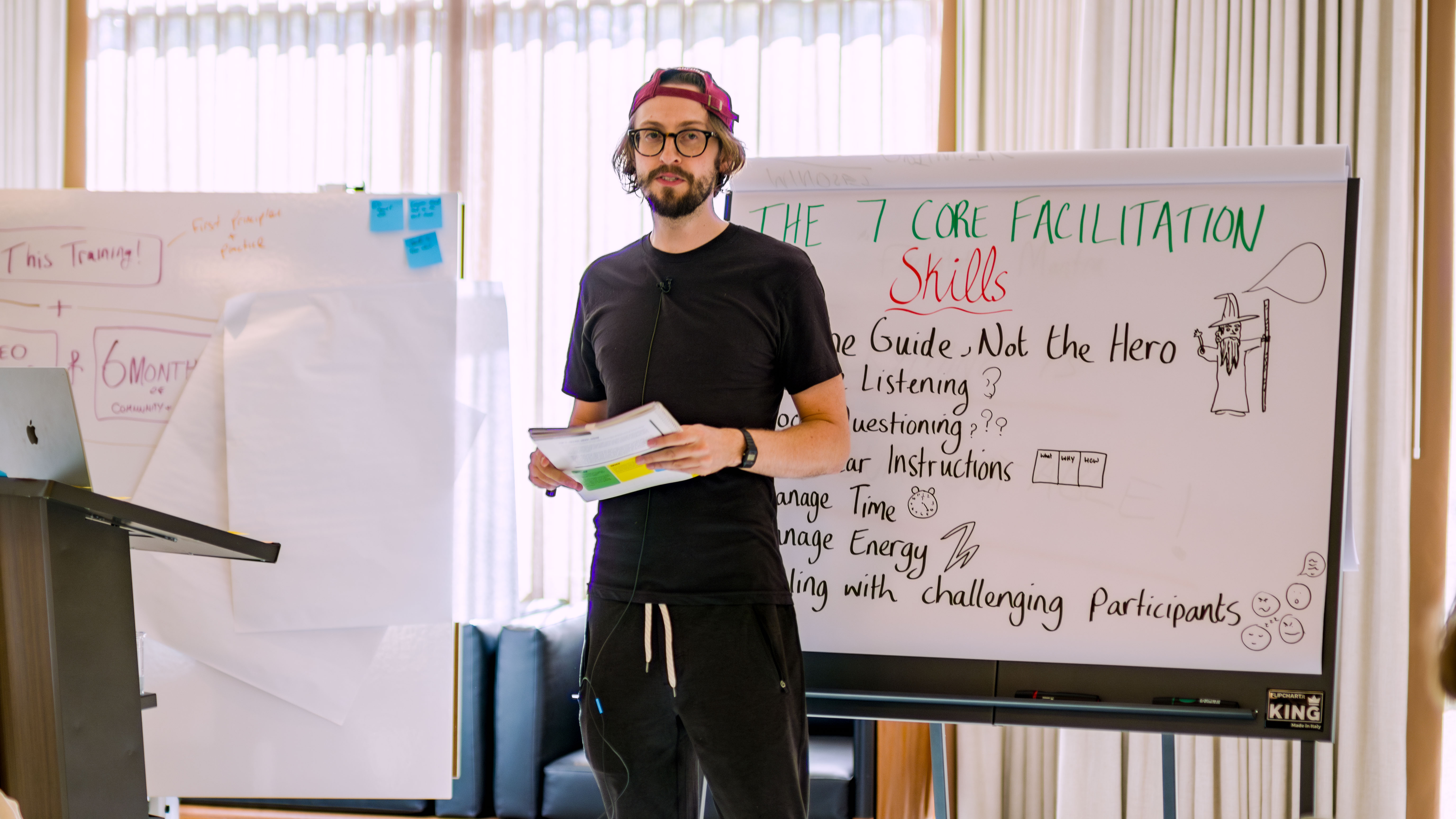 A facilitation trainer holding workshop materials stands in front of a whiteboard listing the 7 core facilitation skills, including listening, questioning, clear instructions, and managing energy