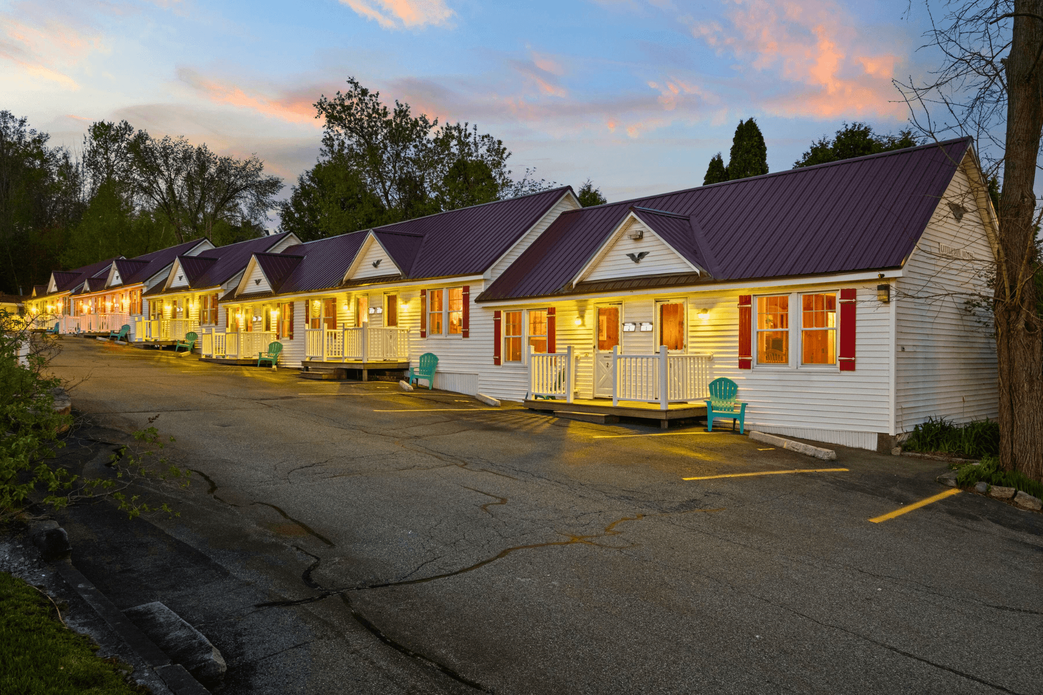 Sunset view of the historic Littleton Motel in New Hampshire, showing a row of white 1940s-style guest rooms with metal roofs, front porches, and Adirondack chairs—part of Lodgism’s top-rated Airbnb offerings.