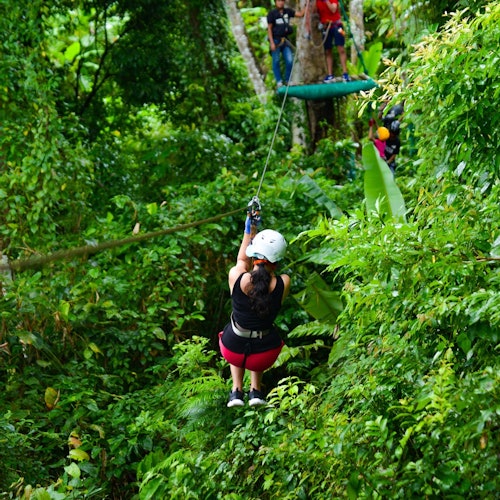 一个人戴着头盔和安全带在茂密的热带森林中滑索。背景中有其他人在平台上等待。