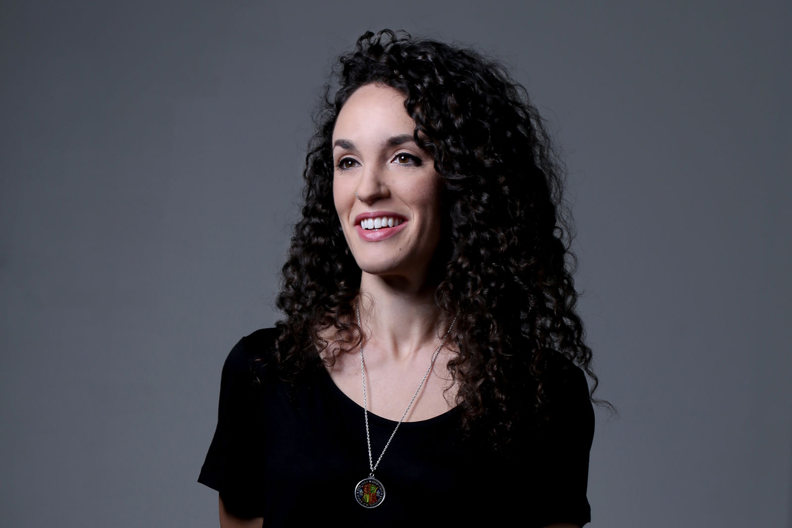 A woman with dark, curly hair wears a black top and a silver necklace with a pendant. She is smiling and looking directly at the camera against a solid gray background.