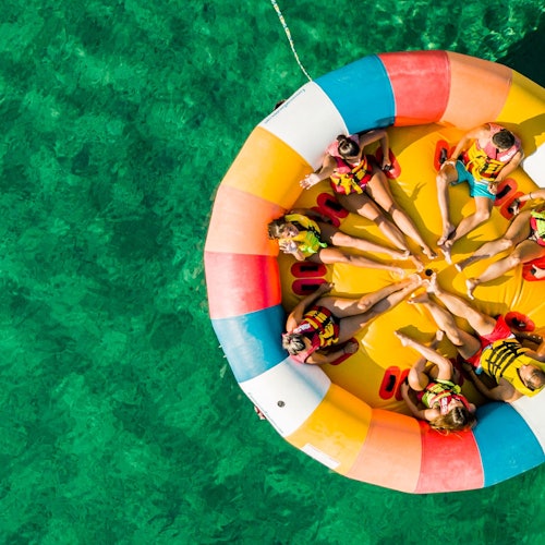 People with life jackets sit on an inflatable colorful raft in clear green water, viewed from above.
