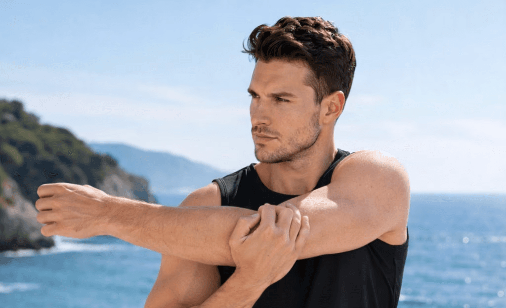 Athletic man in a sleeveless black outfit posing confidently outdoors under a bright blue sky, with a close-up of navy and salmon-colored men’s running shoes in the foreground.