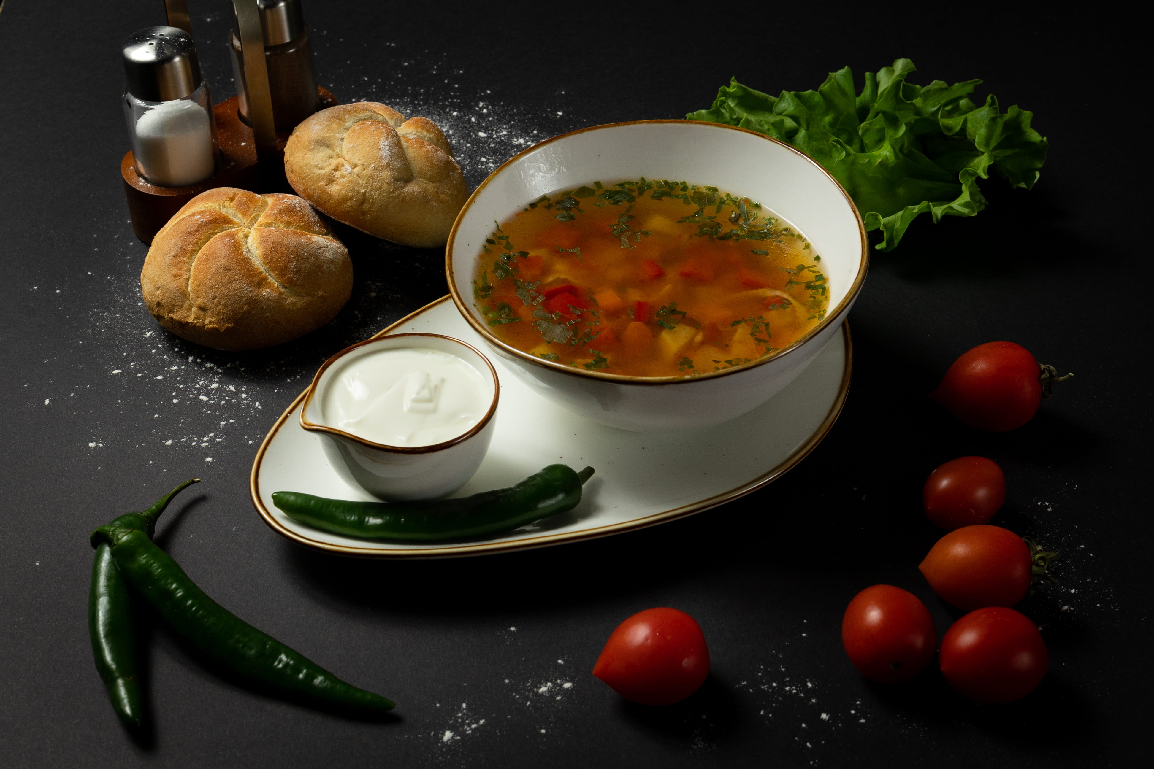 Un bol cu ciorbă de legume presărată cu verdeață, servit pe un platou oval alături de un sos de smântână, ardei iuți verzi, chifle calde și roșii cherry, pe un fundal negru.