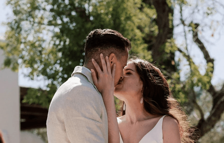 Een bruid en bruidegom delen een romantische kus in een buitenomgeving, omringd door groen en witte decoratie.