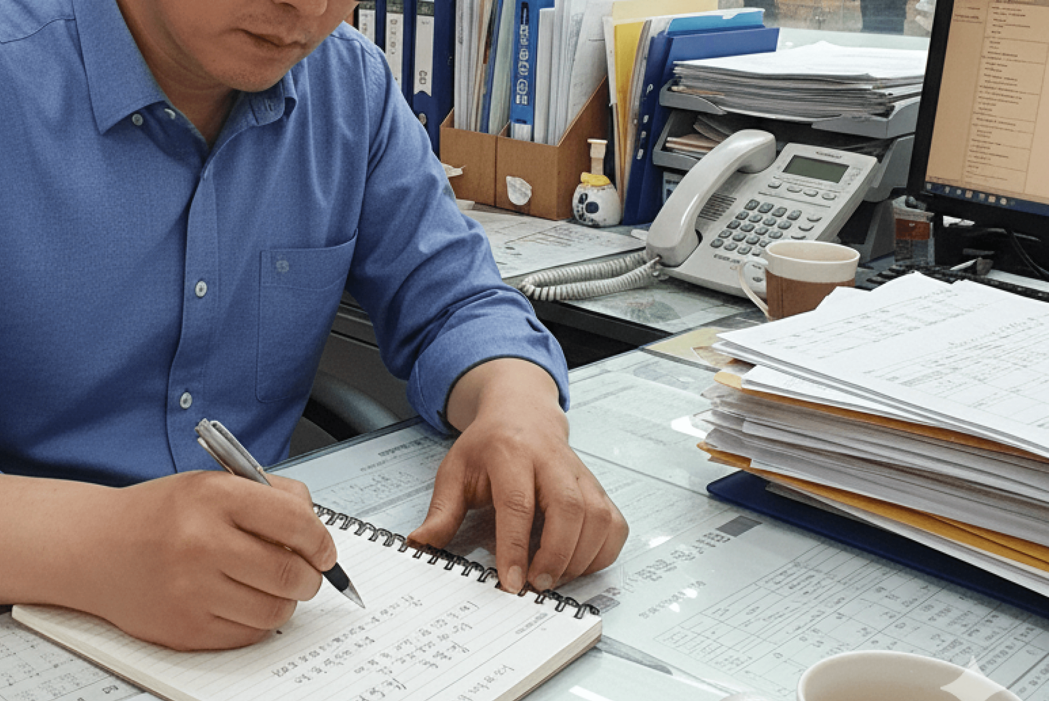 한 남성이 푸른색 셔츠를 입고 사무실 책상에 앉아 줄이 그어진 노트에 검은색 펜으로 무언가를 수기로 작성하고 있는 모습. 책상 위에는 서류 뭉치, 전화기, 종이컵, 컴퓨터 모니터 등이 놓여 있으며 업무에 집중하고 있는 분위기를 보여줍니다.