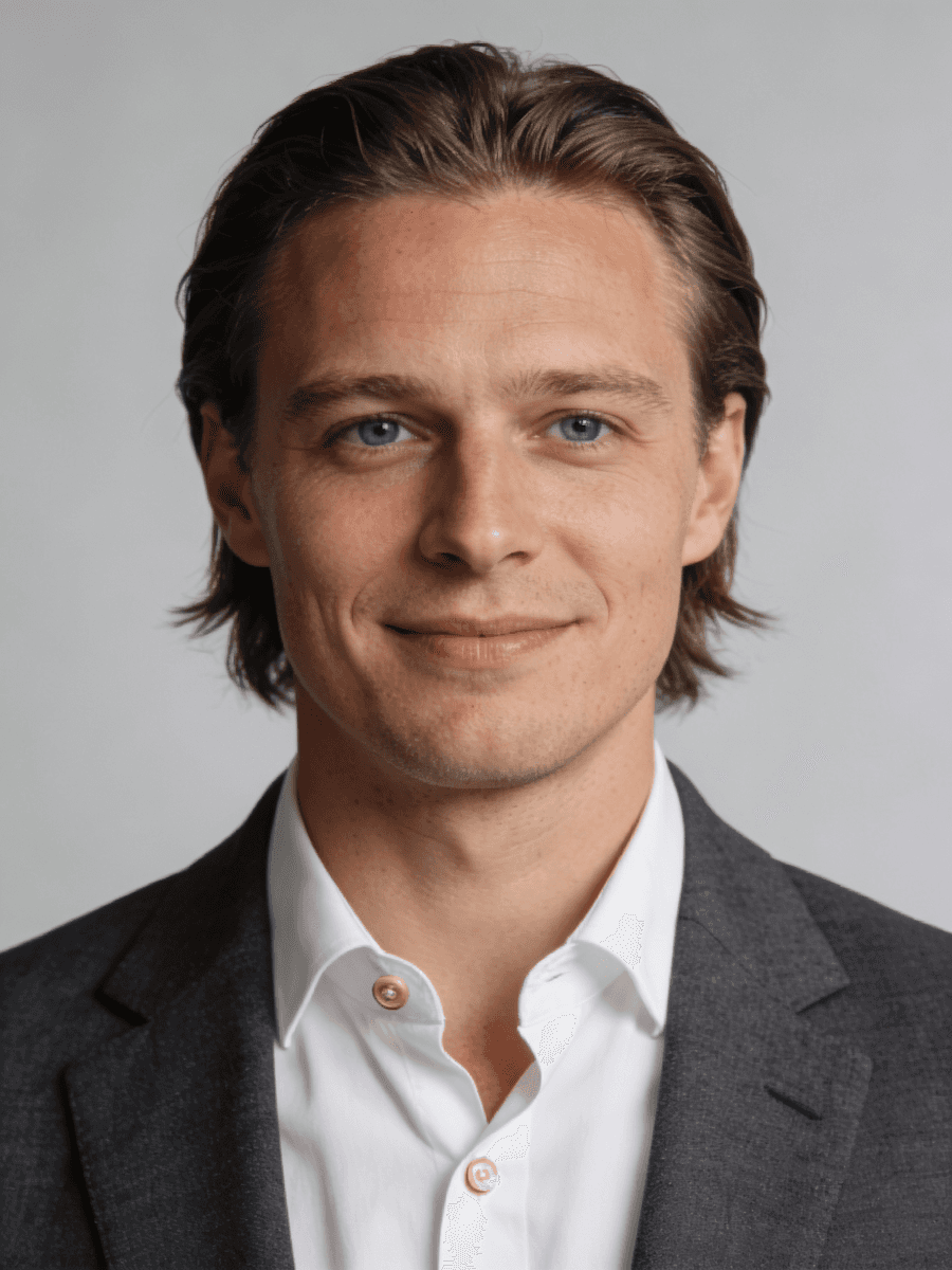 Headshot of a smiling man with brown hair and blue eyes, wearing a dark blazer and white shirt on a grey background.