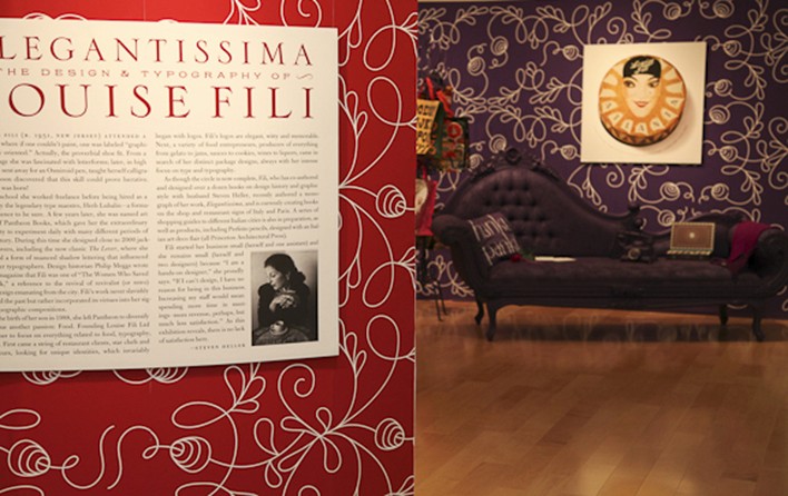 image of a poster with a big red title and two columns of small text under it, and in the back is a couch and a purple wallpaper with a white curved line pattern