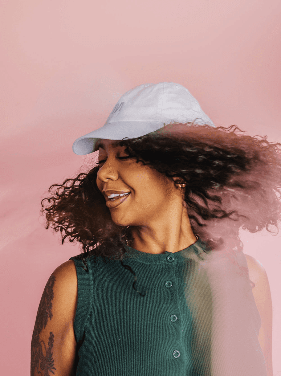 Woman with curly hair wearing a white cap, turning her head mid-motion against a soft pink background.