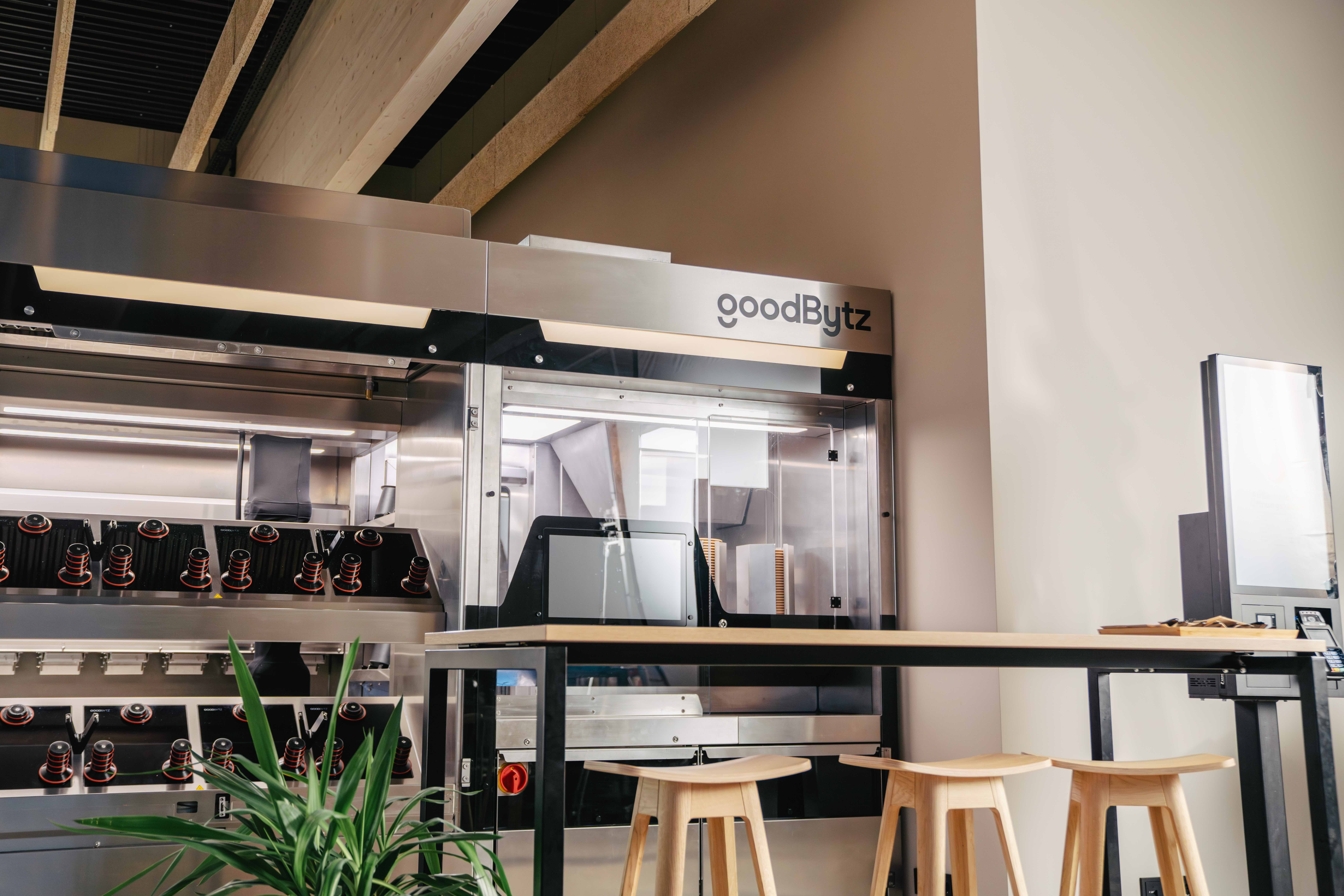 Modern café interior with wooden stools, metal railings, and open kitchen space. Natural light enhances the ambiance.