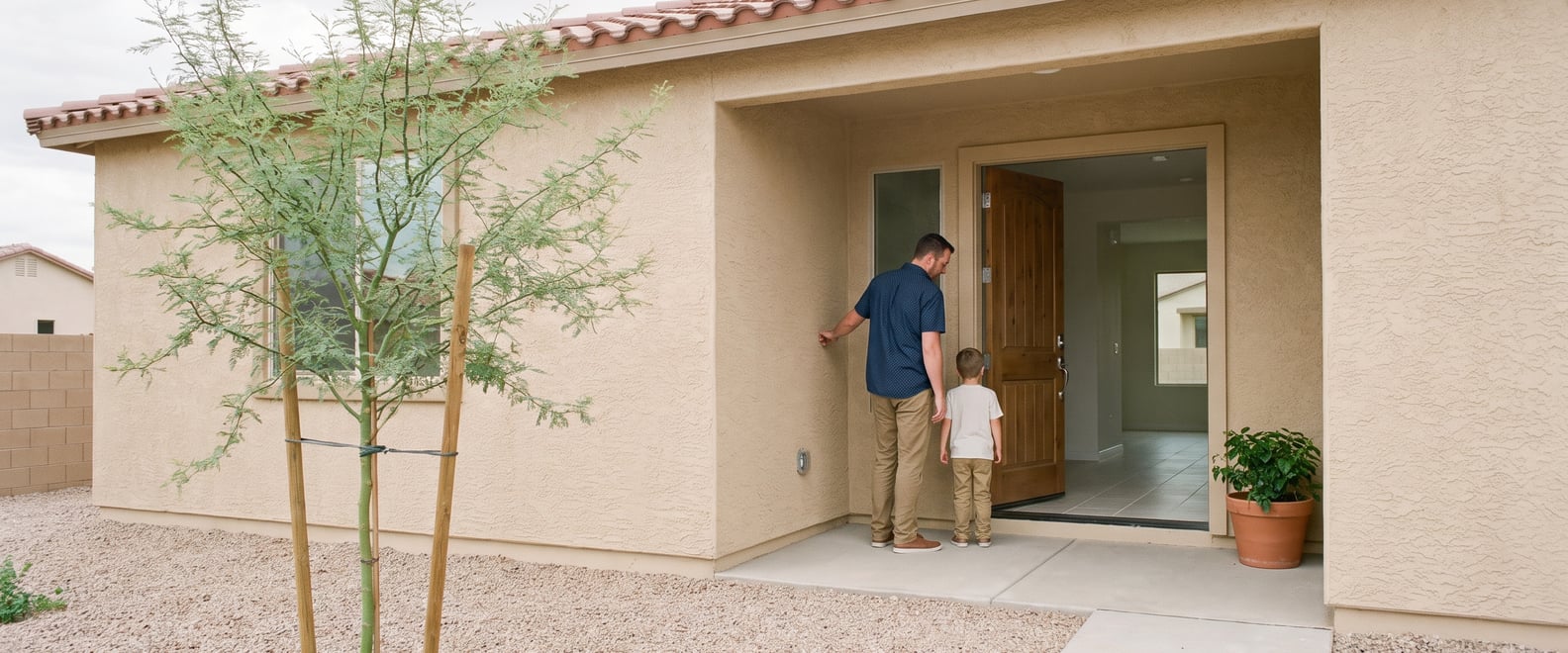 A homeowner and his young son standing on the porch of a newly built Chandler stucco house, open front door, gravel yard and a potted plant
