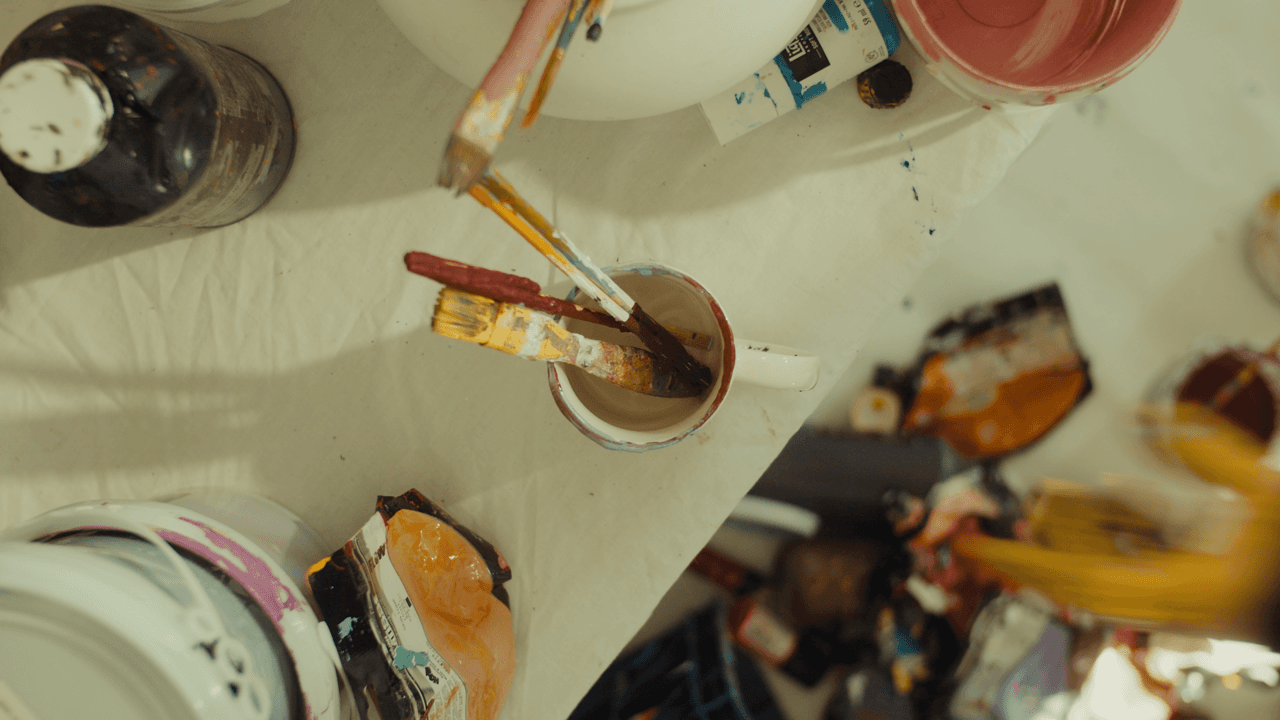 Close-up of paint brushes, tubes of paint, and art tools laid out on a wooden table in the middle of an artistic workspace