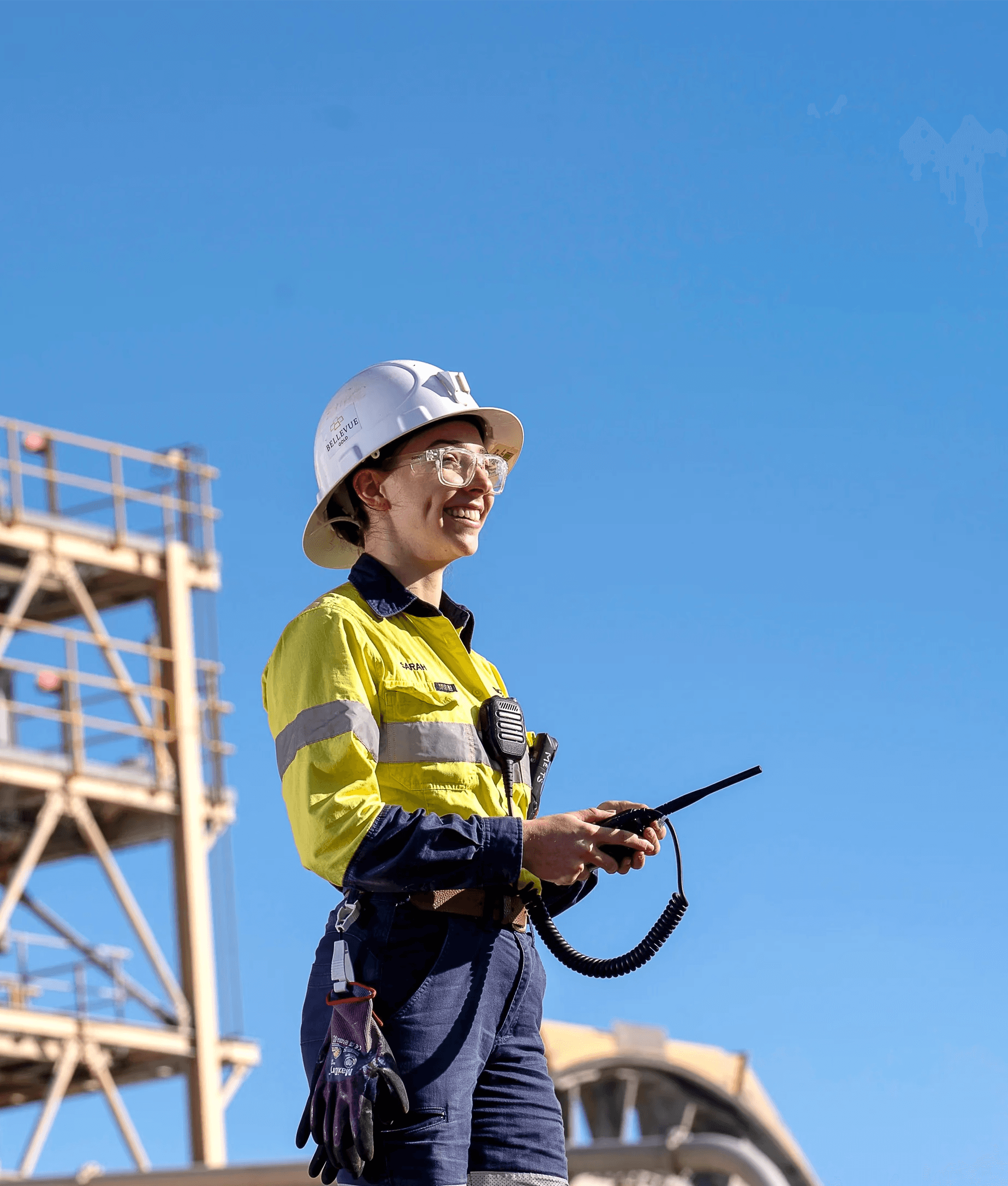A female mining engineer wearing a white hard hat, safety glasses, and high-visibility yellow and navy clothing, smiling and holding a two-way radio under a clear blue sky at Bellevue Gold Mine