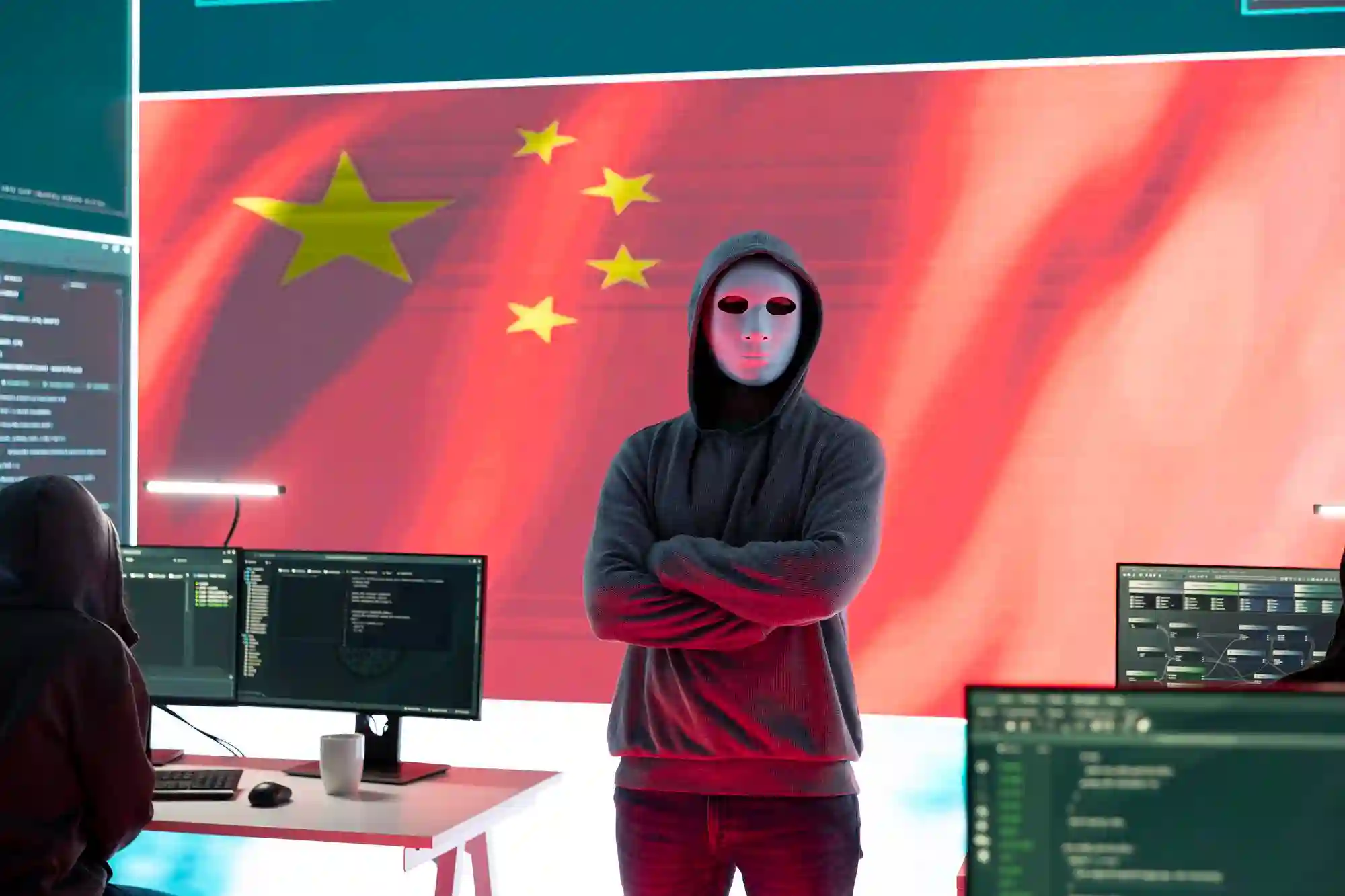 A hooded figure in a mask stands before a Chinese flag and computer monitors, symbolizing state-sponsored cyber espionage.