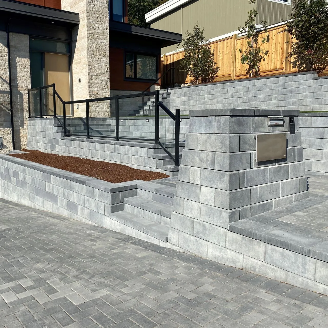 An entrance across levels Gray steps and tiered walls lead from a paved approach to a modern home, with black railings and raised planting beds.