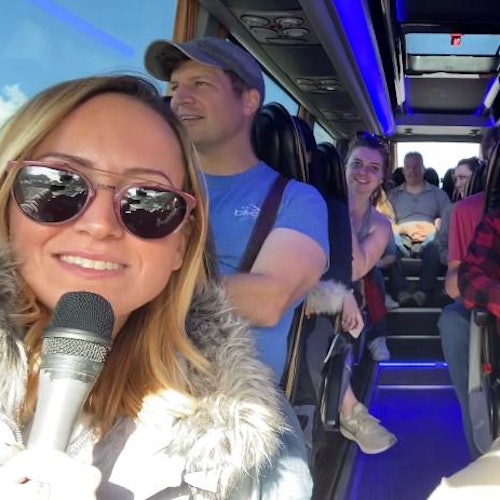 A woman holds a microphone and smiles at the camera inside a bus with other passengers seated behind her, smiling and relaxed.