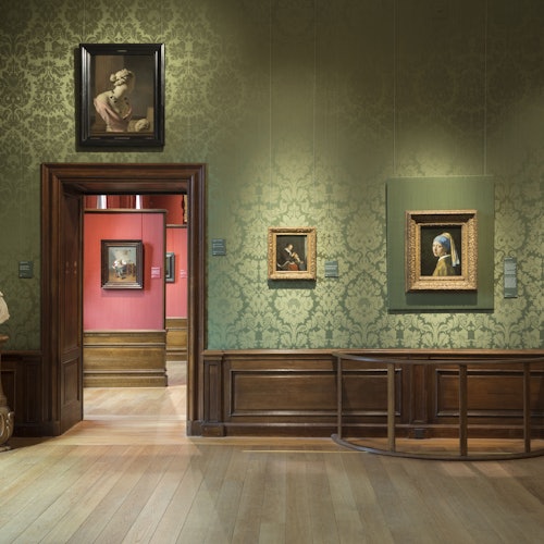 博物館的房間有綠色的牆紙、木地板和框架畫作。一扇門通往另一個紅色牆壁的畫廊。