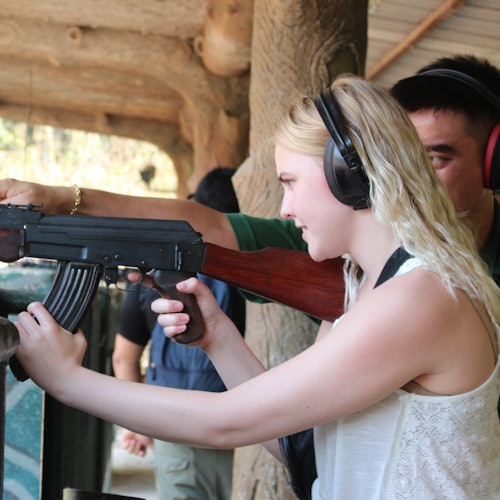 Seorang wanita berambut pirang mengenakan pelindung telinga mengarahkan senapan di lapangan tembak, dibantu oleh seorang pria di latar belakang.