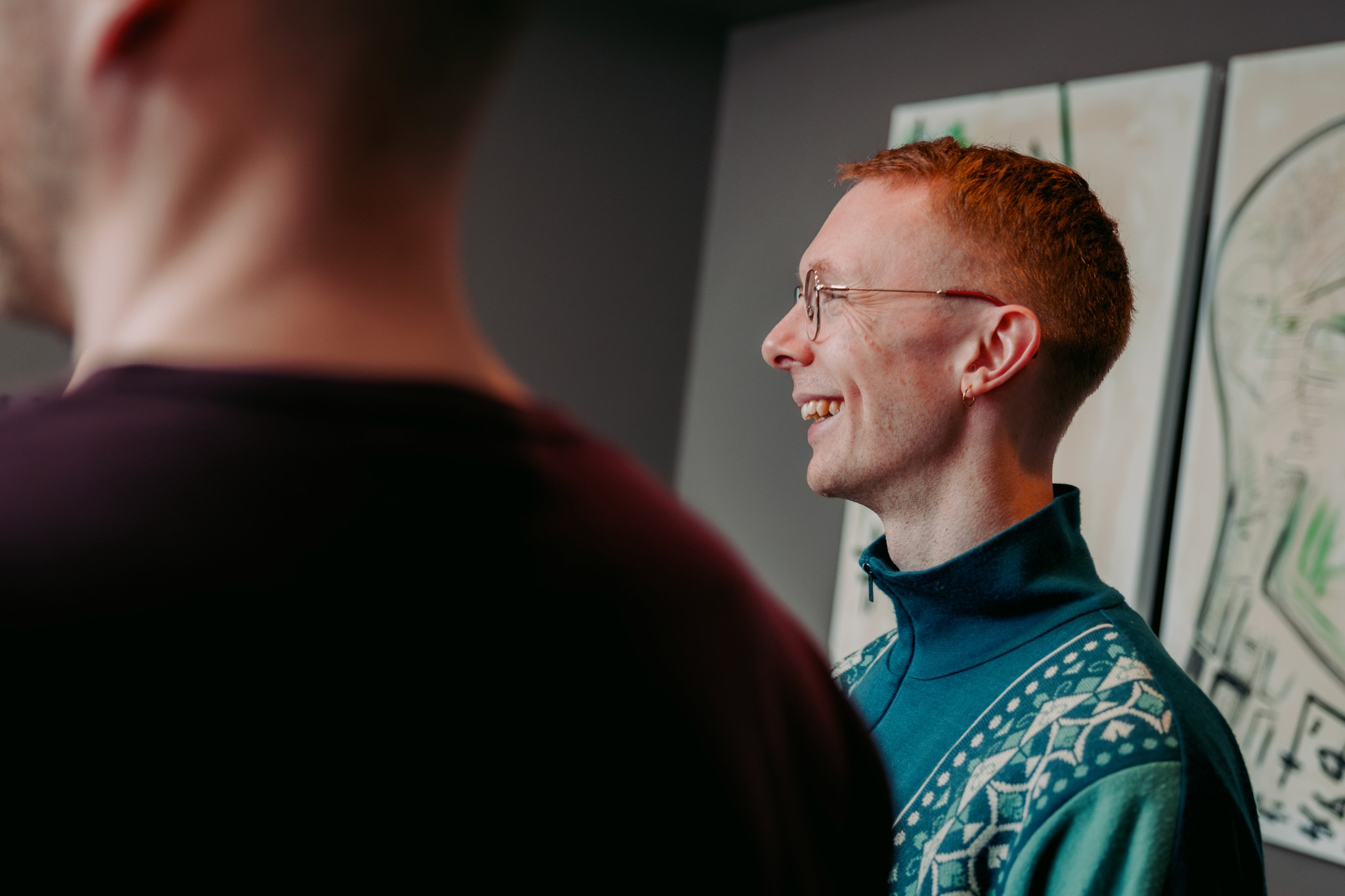 Two people are in a room with modern decor; one person in the foreground faces away, while another with red hair and glasses smiles, wearing a teal patterned sweater.