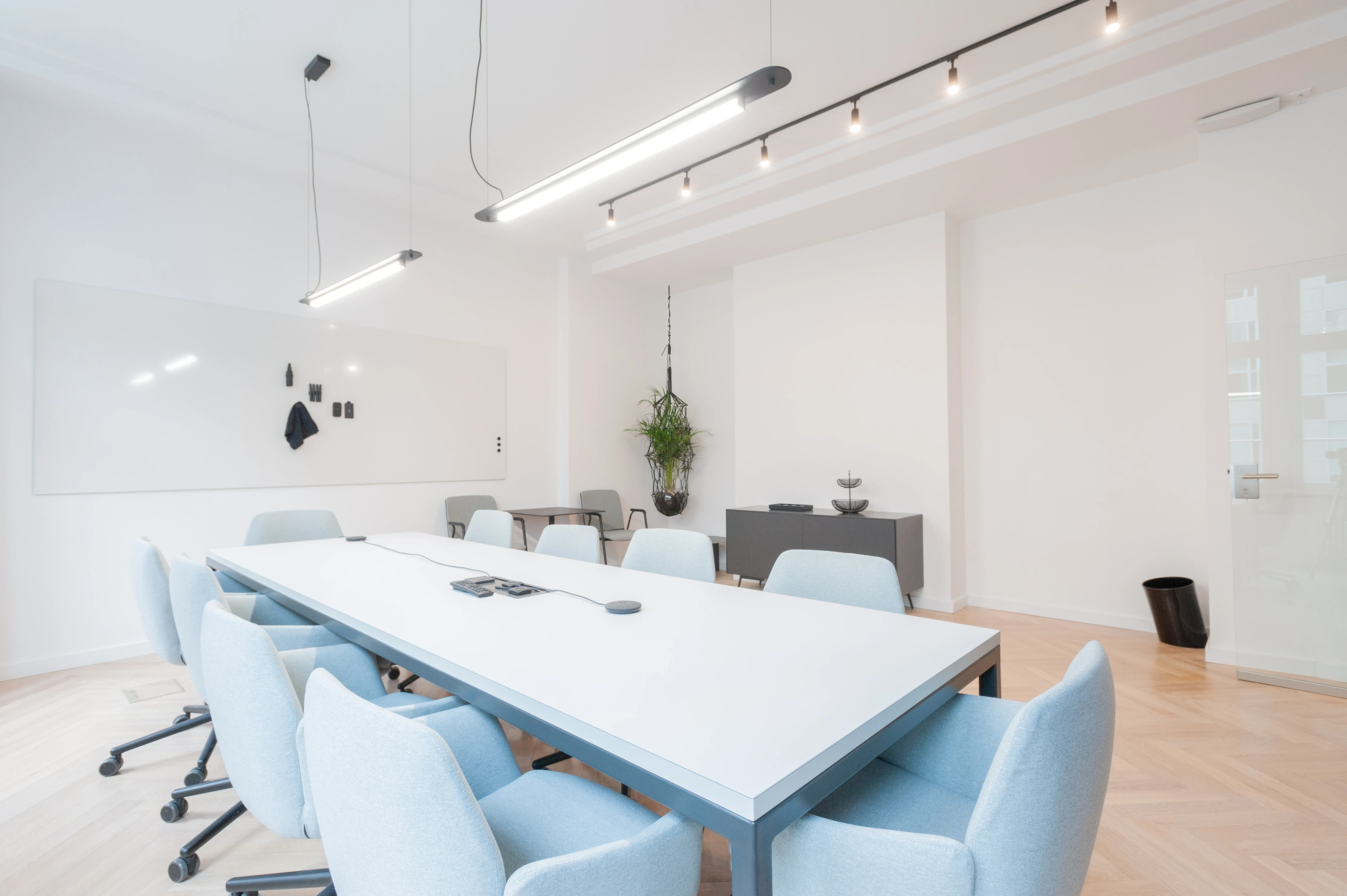 Bright meeting room at AI START-UP BERLIN with a long white table, pale blue upholstered swivel chairs, a whiteboard wall, a hanging plant and black linear pendant lights on a track.