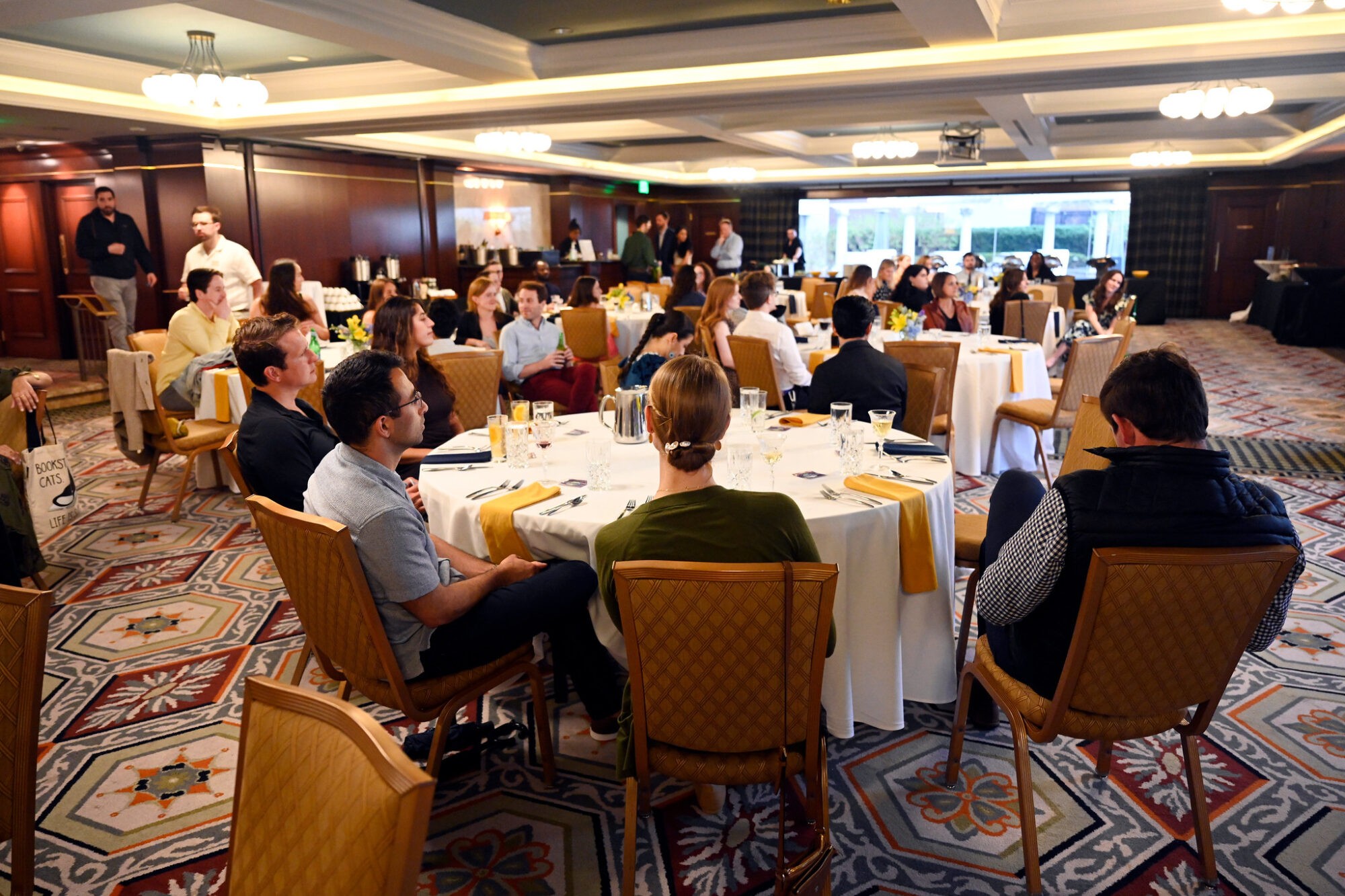 A group of people seated around tables in a conference room, engaged in a presentation or discussion.