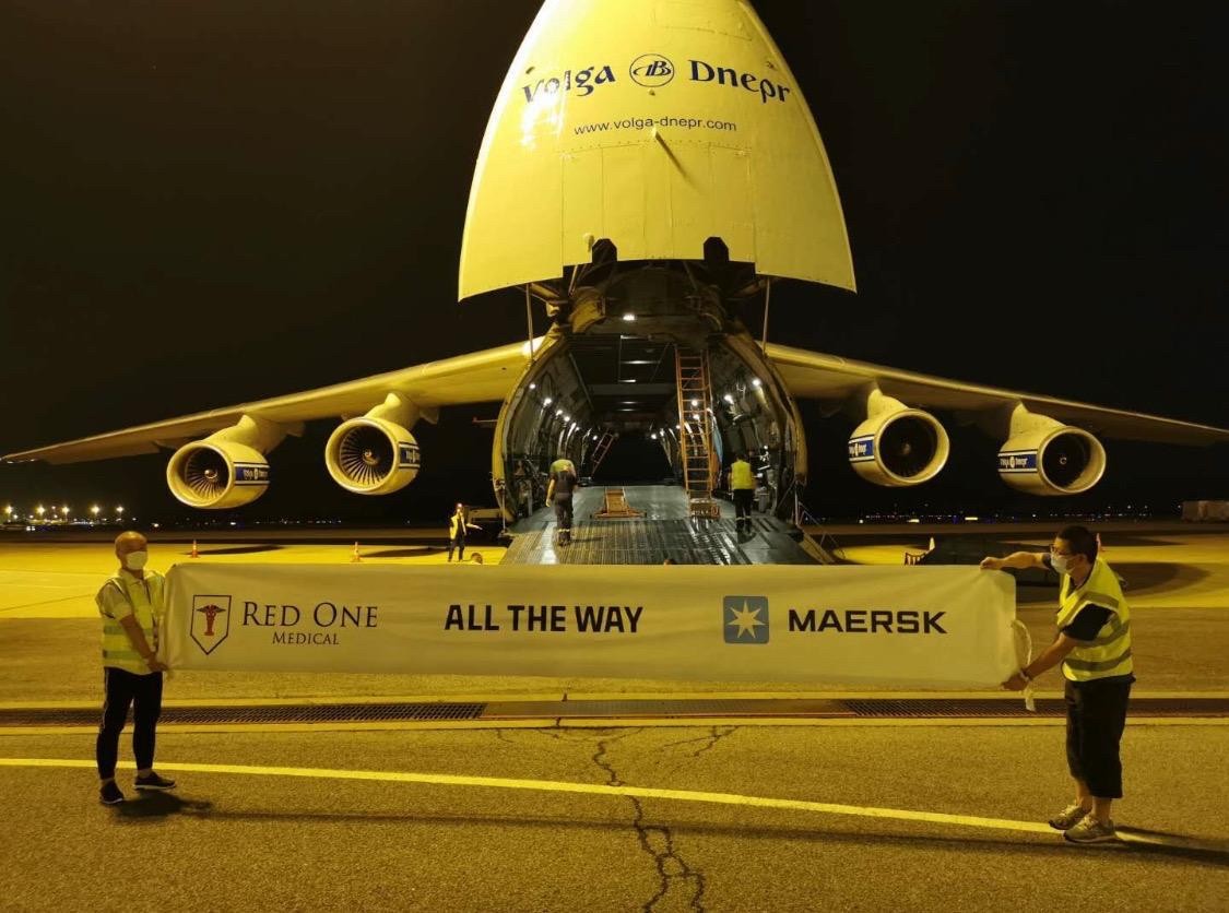 A large cargo aircraft on the tarmac, with two people standing in front, holding a sign. Nighttime setting.