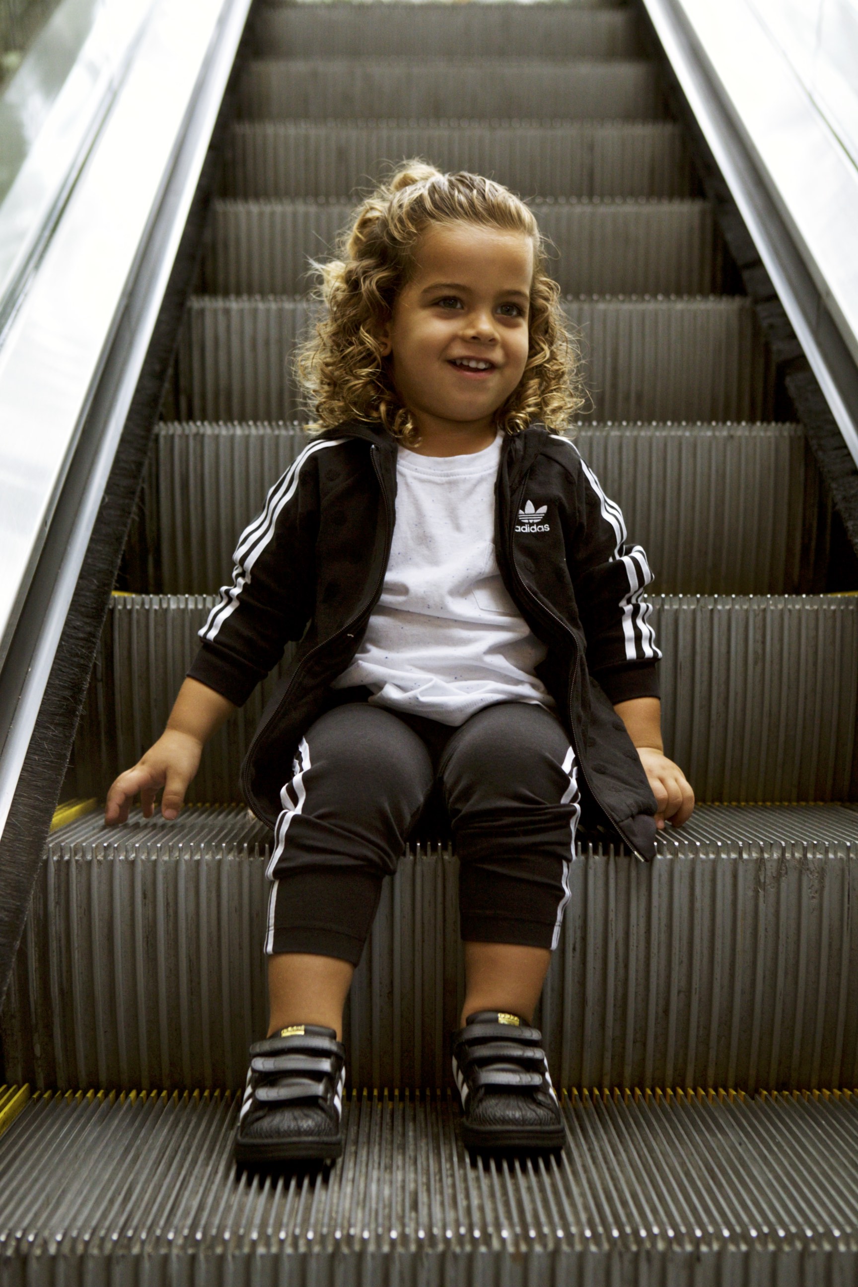 Toddler in a black adidas tracksuit sitting on an escalator step, smiling.