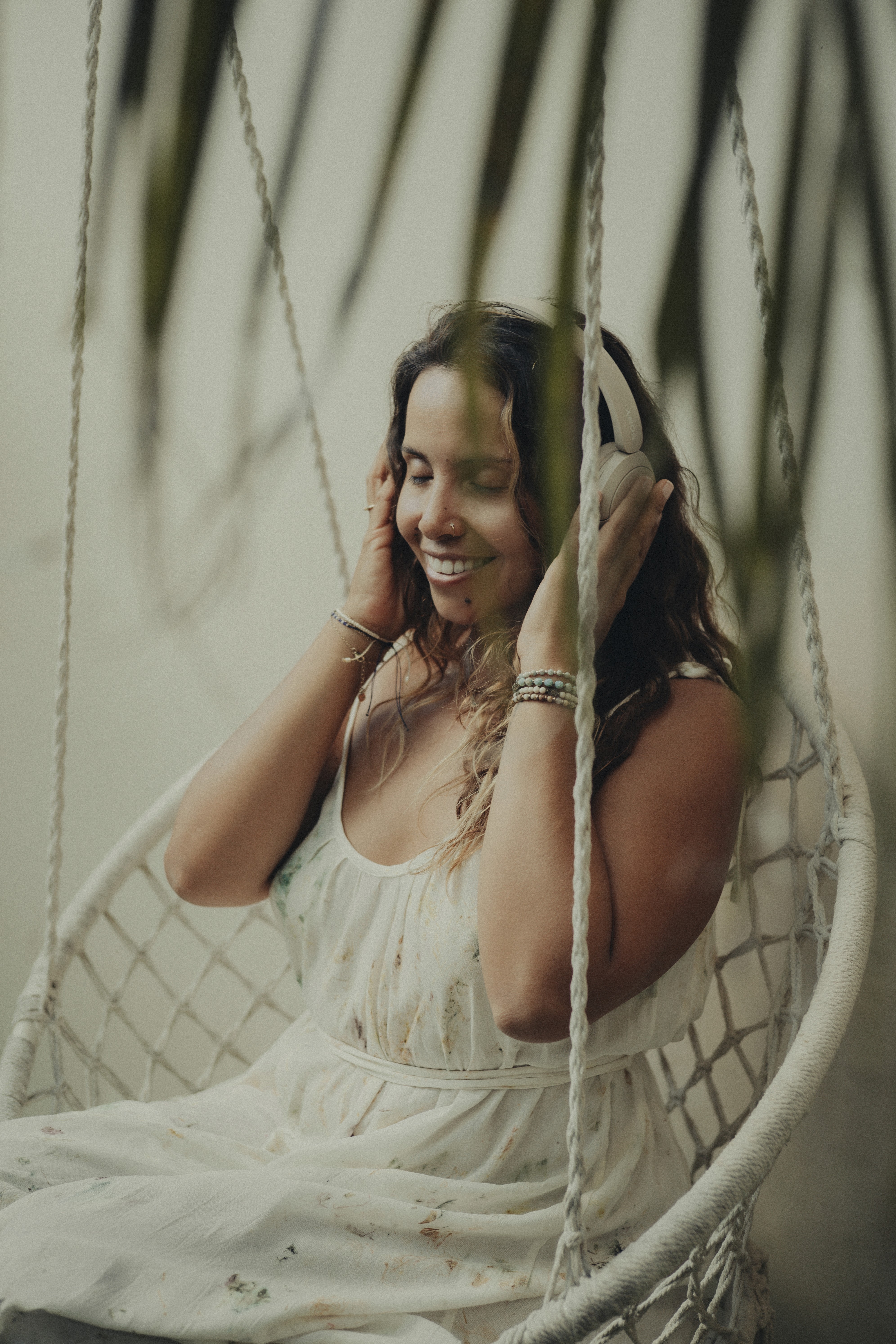 Smiling girl with closed eyes, wearing headphones and relaxing on a hanging chair, surrounded by green plants