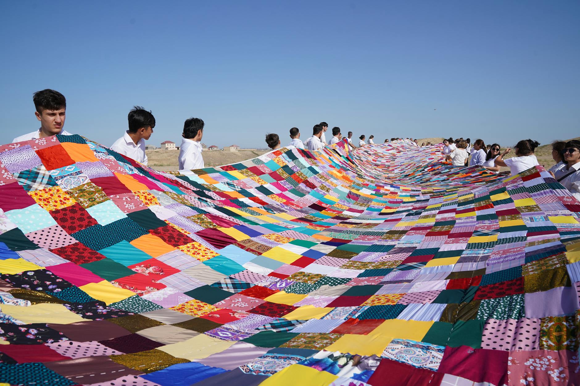A large multicolored patchwork quilt is held by a group of people outdoors on a sunny day.