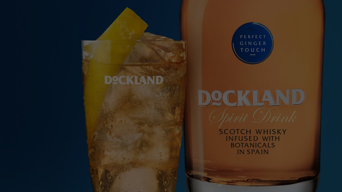 A glass of iced Scotch whisky with a lemon slice is beside a Dockland Spirit Drink bottle. The label reads "infused with botanicals in Spain."