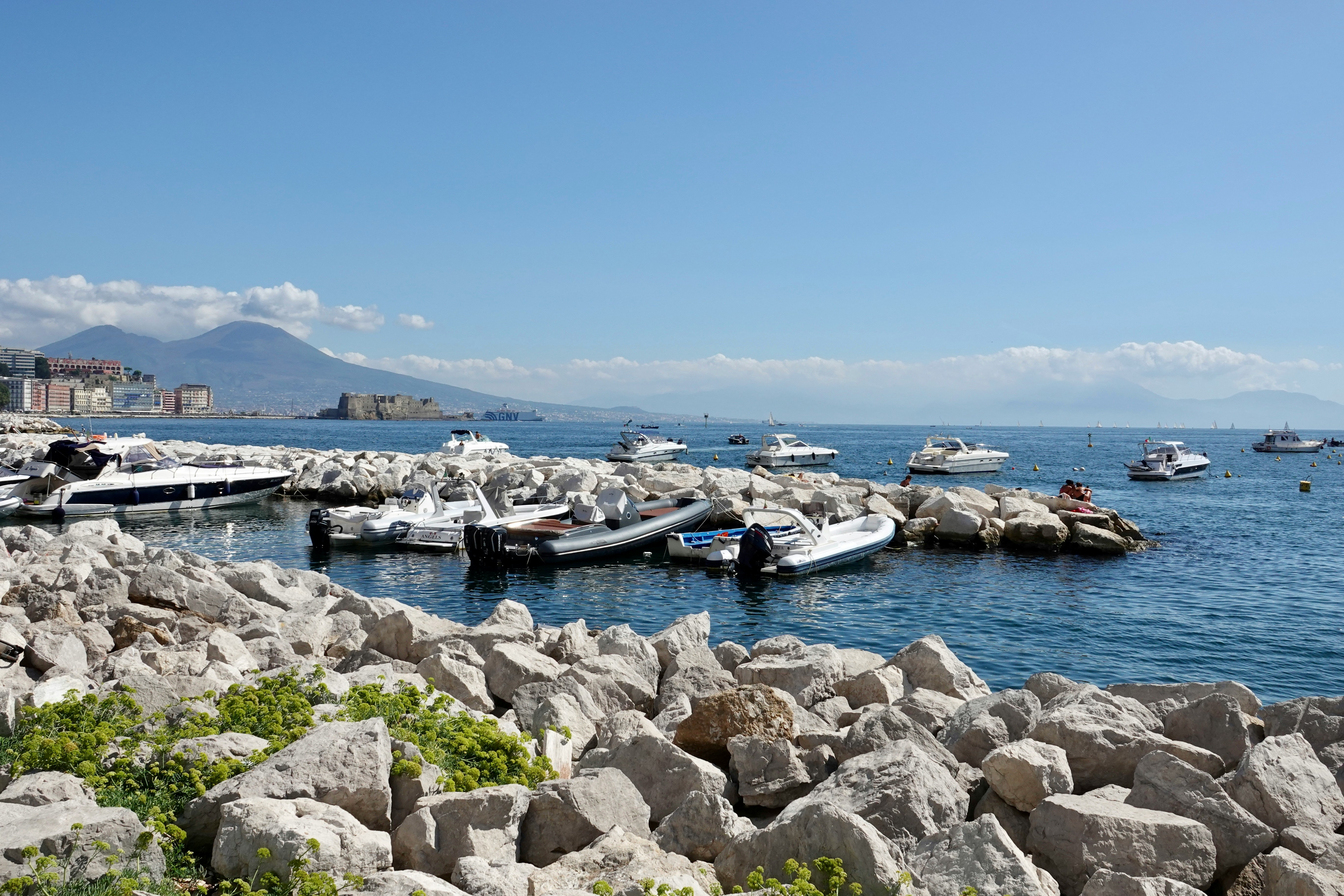 Una vista panoramica del lungomare di Napoli, Italia, con piccole barche ormeggiate lungo le coste rocciose, una vegetazione vibrante in primo piano e il Vesuvio sullo sfondo sotto un cielo azzurro limpido.