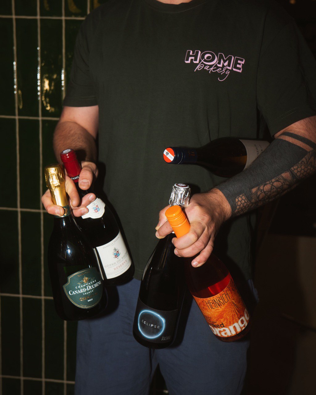 A person holding multiple bottles of assorted wine and champagne, each with distinct labels, standing against a green tiled background, showcasing a diverse selection of beverages.