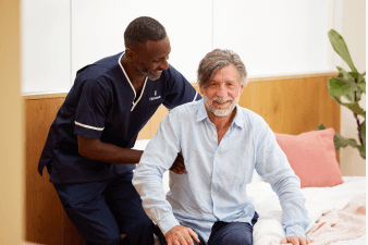 A male nurse in dark scrubs sitting with an older man in a relaxed indoor setting, engaged in conversation or a care assessment