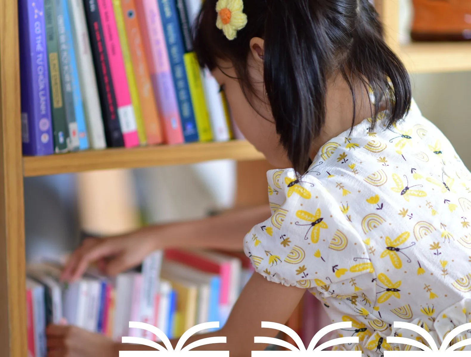 A young girl with pigtails looking through library books with three white book elements in the bottom white corner.